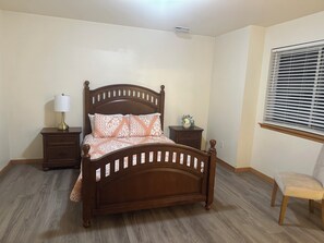 A bedroom with a wooden bed, nightstands, a lamp, a chair, and a window with blinds.