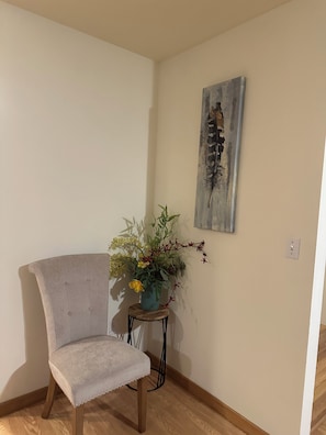 Une pièce d’angle avec une chaise de couleur claire, une petite table sur laquelle est posé un vase de fleurs et une œuvre d’art accrochée au mur.