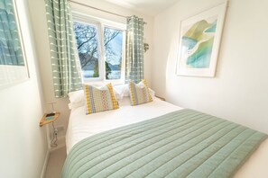 Ein Schlafzimmer mit Bett, Kissen, einem Nachttisch, einem Fenster mit Vorhängen und einem Bild an der Wand.