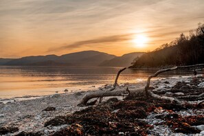 Eine Seenlandschaft bei Sonnenuntergang, mit Bergen im Hintergrund und einem steinigen Ufer mit Treibholz.