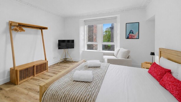 A bedroom with a bed, a wooden bench, a TV, a window, and a painting.
