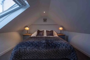 A bedroom with a sloped ceiling, a bed with a dark quilt, two bedside lamps, and a small wall-mounted decoration. - Fife Cottage between St Andrews and Dundee (Fife)