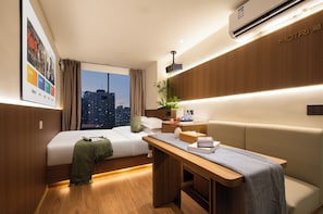 Ein modernes Hotelzimmer mit Bett, Schreibtisch, Sofa, einer Topfpflanze und Blick auf die Stadt.