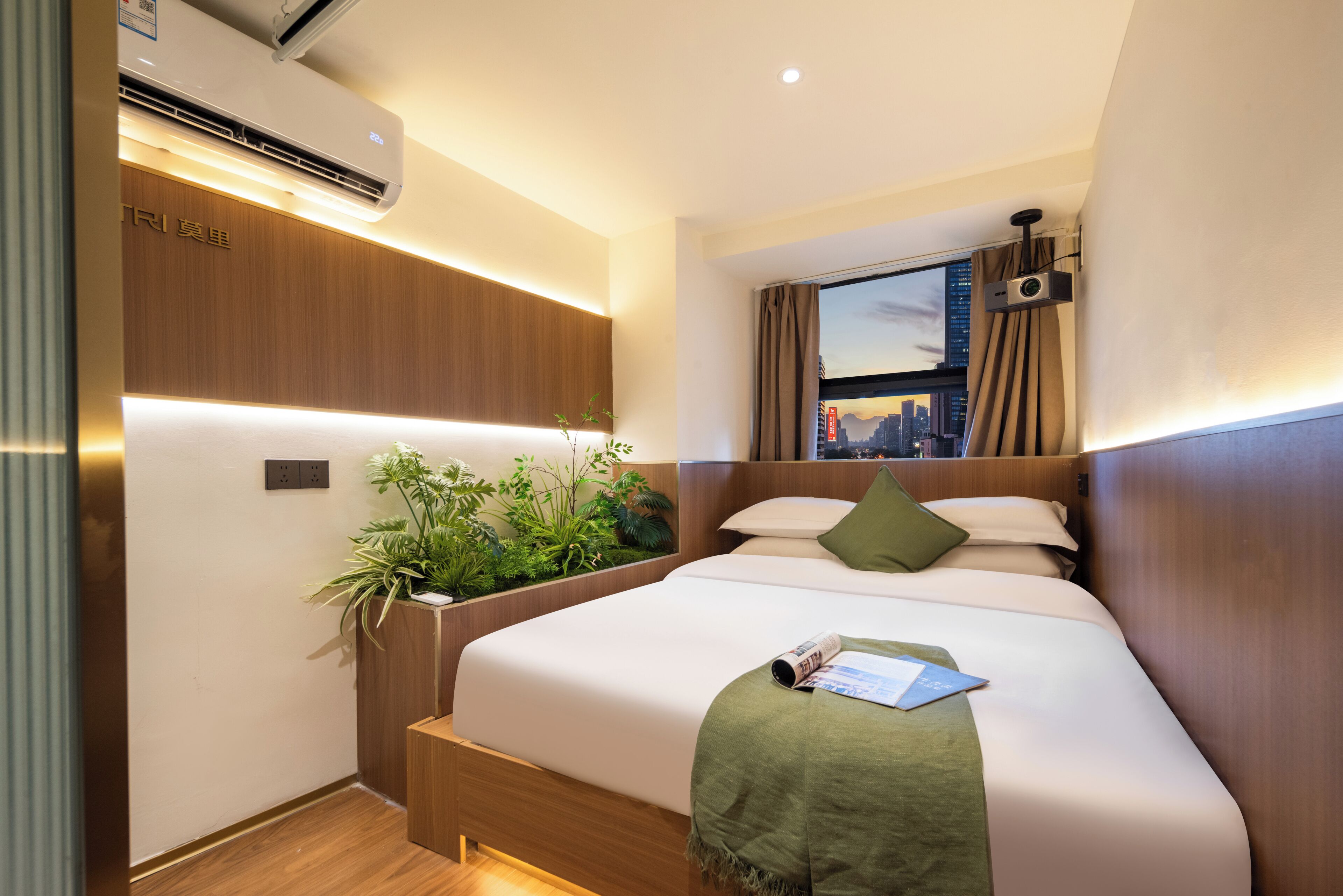 A hotel room with a bed, a window with curtains, an air conditioner, a plant, and a cityscape view.