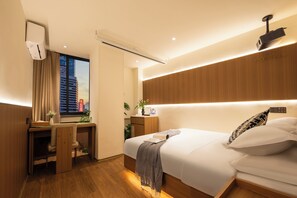 Quarto de hotel moderno com escrivaninha de madeira, cama com roupa branca, mesa de cabeceira com abajur e vista para a paisagem urbana.