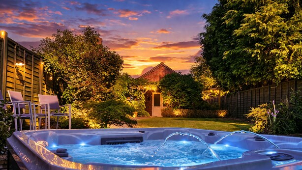 A hot tub with water jets, surrounded by a wooden deck, chairs, and a brick house in the background. - Lilac Cottage (Henley-In-Arden)