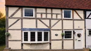 A traditional half-timbered house with a stone foundation, multiple windows, and a white door. - Lilac Cottage (Henley-In-Arden)