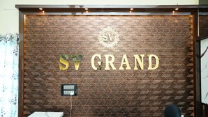 A reception desk with a herringbone patterned wall, a logo with 'SV' and 'GRAND' text, and a black chair. - SV Grand (Kakinada)