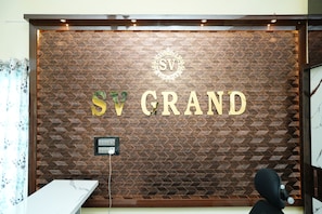 Un mostrador de recepción con un revestimiento de pared tipo espiga, un logotipo con el texto 'SV' y 'GRAND', y una silla negra.