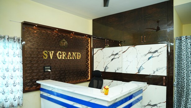 A reception desk with a marble top and blue accents, a patterned wall behind it, and a dark wooden cabinet above. - SV Grand (Kakinada)
