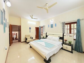 A bedroom with a bed, a nightstand, a chair, a ceiling fan, and a window with curtains. - Sunny Brooks 6BHK Spacez Villa with Outdoor Patio & Garden (Bengaluru)
