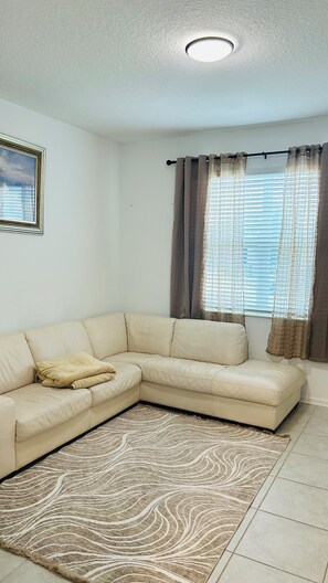A living room with a beige sectional sofa, a patterned rug, a ceiling light, and a window with curtains. - Cosy 1-bedroom house with AC in Haines City (Haines City)