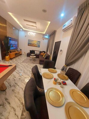 A modern living room with a flat-screen TV, a sofa, a dining table set for four, and a marble floor. - 3-bed apmt - Lekki - AC, Swimming Pool, Gym, Workstn, PS5, 24/7 ElectricSecurity (Lekki)