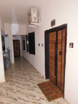 Un couloir doté d’un sol en marbre, une porte en bois portant l’inscription « B11 », et un petit appareil de climatisation fixé au mur.