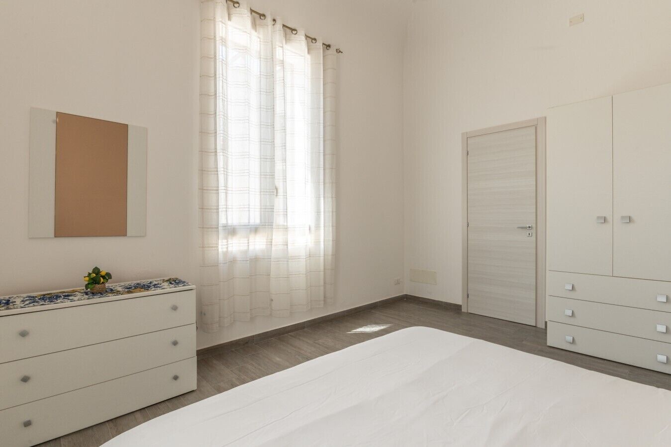 Une chambre à coucher avec un lit, une commode, une armoire, un miroir et une fenêtre avec des rideaux.