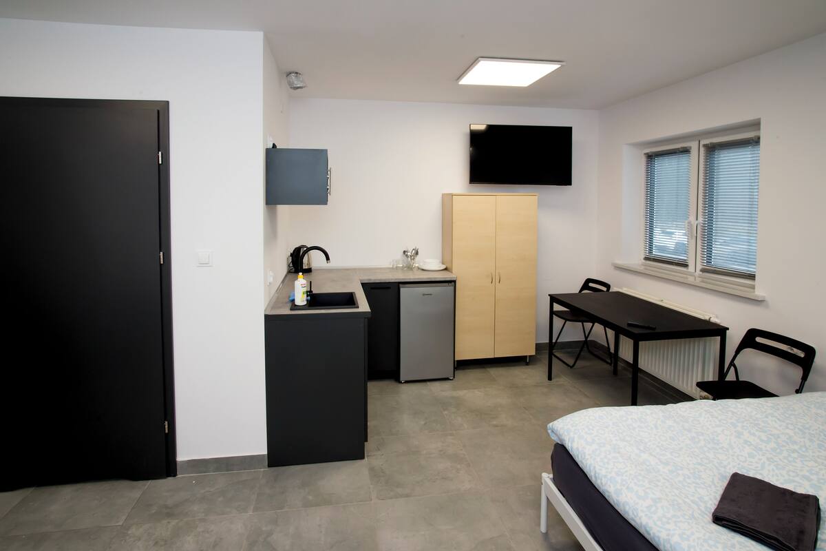 Une pièce de taille modeste comprenant un lit, un bureau, une petite kitchenette, un téléviseur et une fenêtre.
