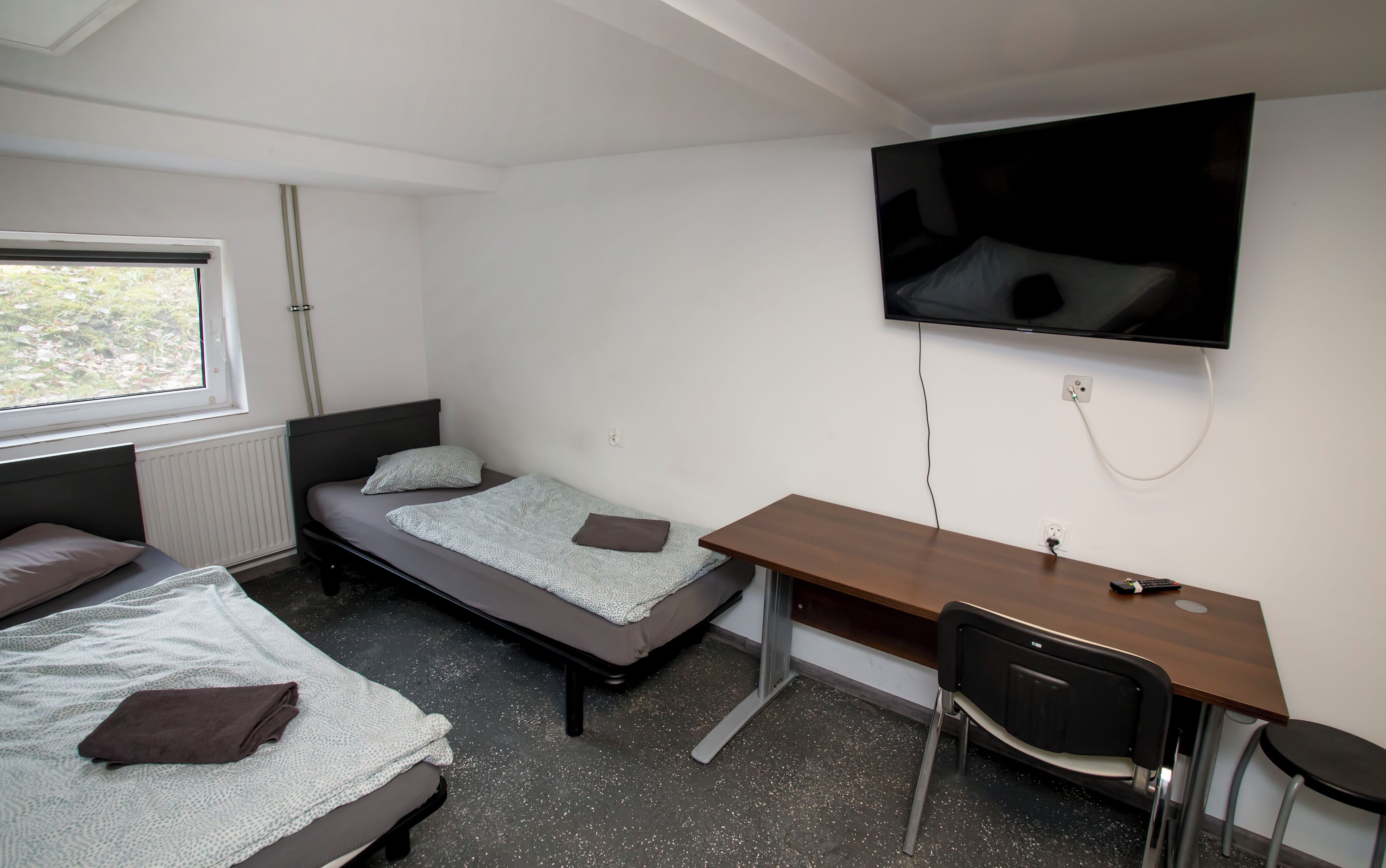 Una habitación tipo dormitorio con una cama, un escritorio, una silla, un televisor y una ventana.