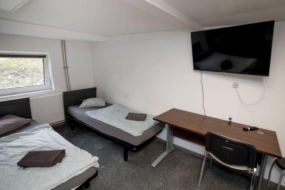 Une chambre de type dortoir, équipée d’un lit, d’un bureau, d’une chaise, d’un téléviseur et d’une fenêtre.