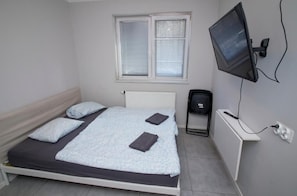 A single bed with a white headboard, a window with blinds, a television mounted on the wall, and a small bedside table with a power outlet.