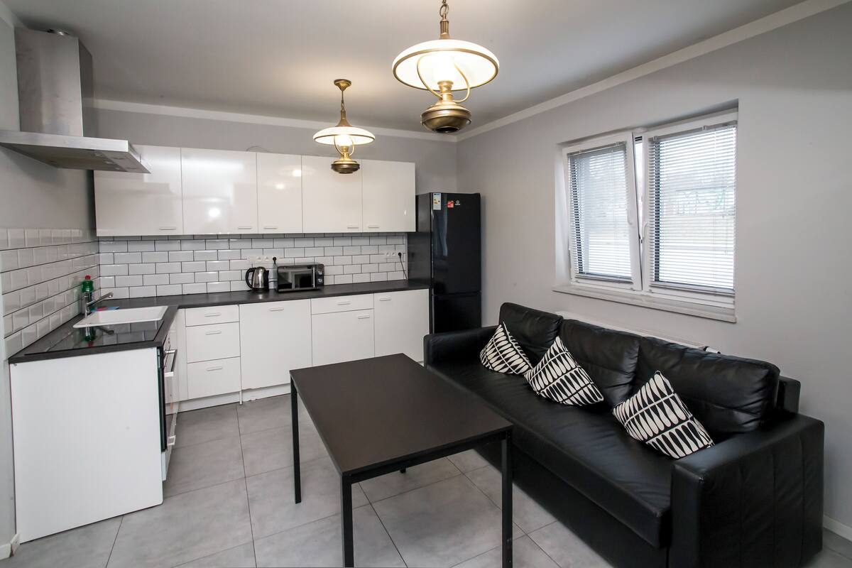 Une cuisine moderne avec des meubles blancs, un réfrigérateur noir, une table à manger et un canapé en cuir noir agrémenté de coussins à motifs.