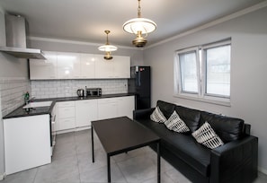 A modern kitchen with white cabinets, a black refrigerator, a dining table, and a black leather sofa with patterned cushions.