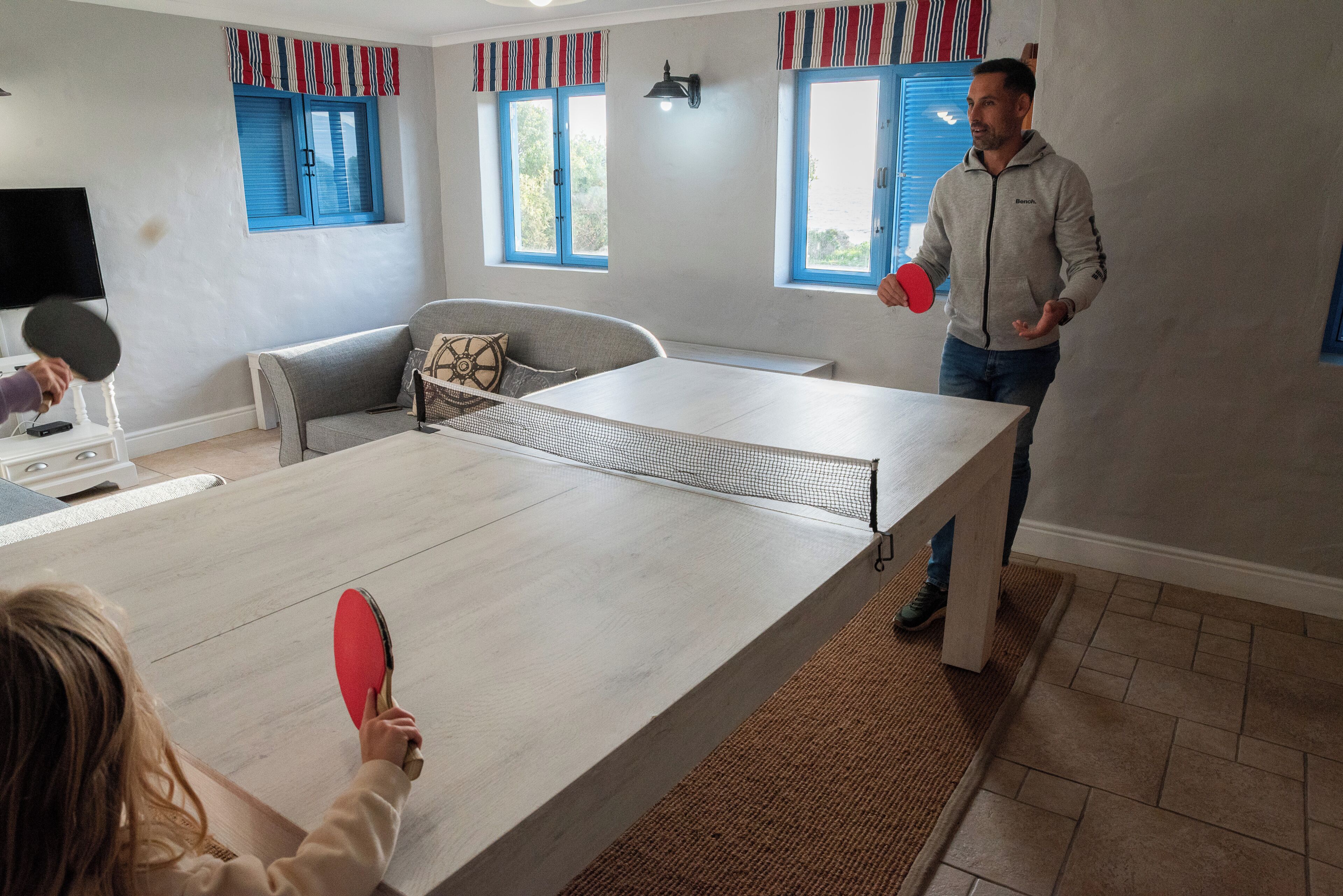 A ping pong table in a room with a sofa, a television, and windows with blue shutters.