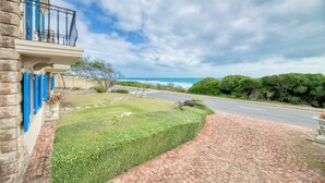 A stone building with blue shutters, a paved driveway, and a view of the ocean. - Casa Balena - The Seafront Whale House Experience (Gansbaai)