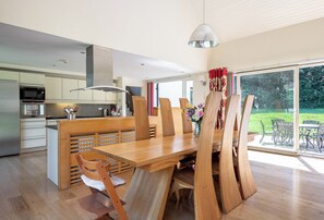 Une cuisine moderne dotée d’un four et d’un micro-ondes encastrés, un coin repas avec des chaises et une table en bois, et une grande porte-fenêtre coulissante donnant sur un jardin.