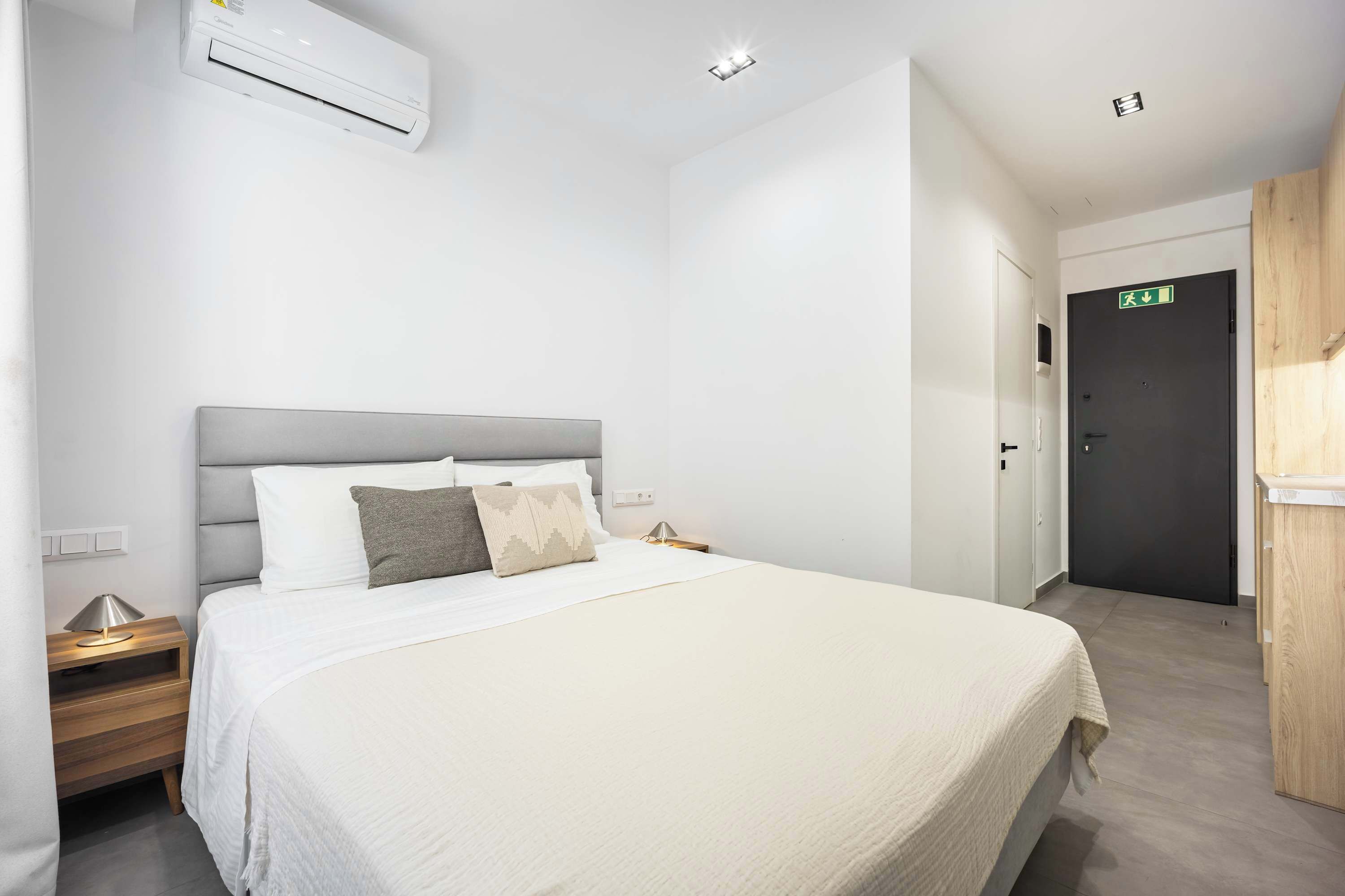 Una camera d'albergo con un letto, comodini, un condizionatore d'aria, una porta con il segnale di uscita e un armadietto in legno.