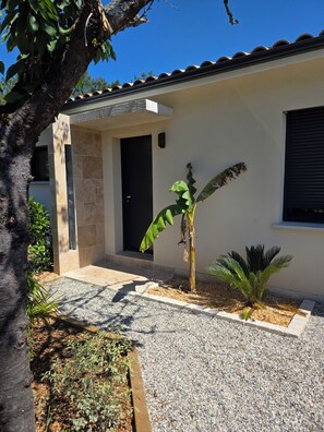 A modern building with a tiled roof, a door, and a small garden area with gravel, a banana tree, and a palm plant. - Summer villa. - Illuminated garden & Brazier (Lesparre-Médoc)