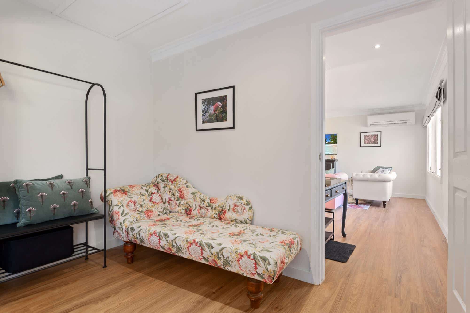 A room with a floral sofa, a small table with a lamp, and a view into another room with a white chair and a desk.