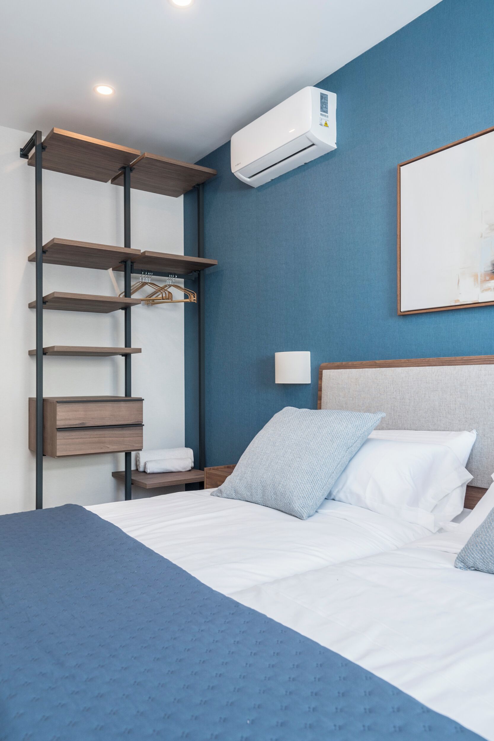 A hotel room with a bed, a wall-mounted air conditioner, a shelf unit, and a painting on the wall.