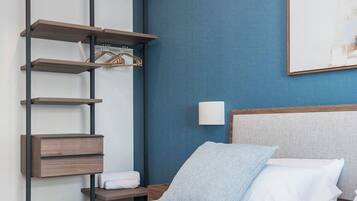 A hotel room with a bed, a wall-mounted air conditioner, a shelf unit, and a painting on the wall.