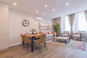 A modern living room with a dining area, a sofa, two armchairs, a coffee table, a clock, and a large window with curtains. - Apartamentos Dúplex Príncipe de Canalejas (Madrid)