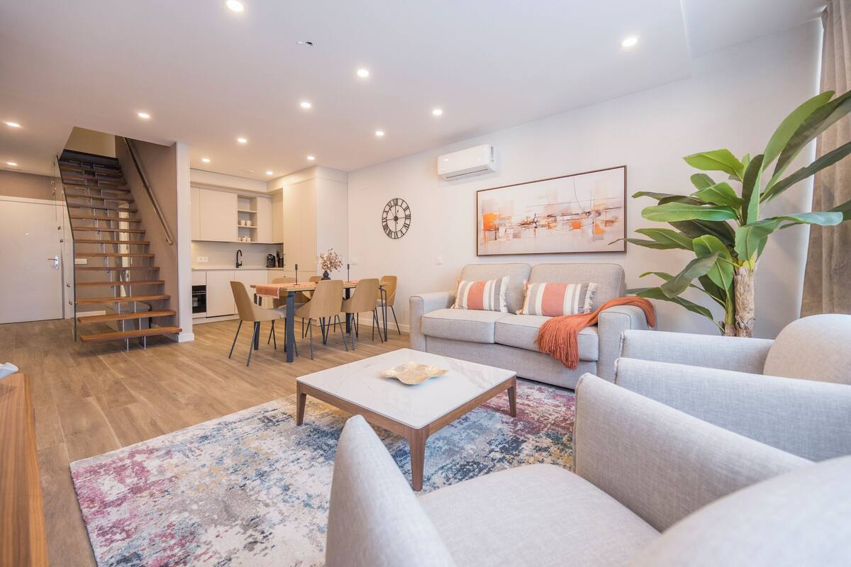 Una sala de estar moderna con sofá, mesa de centro y sillones, una escalera que lleva a una cocina y un comedor.