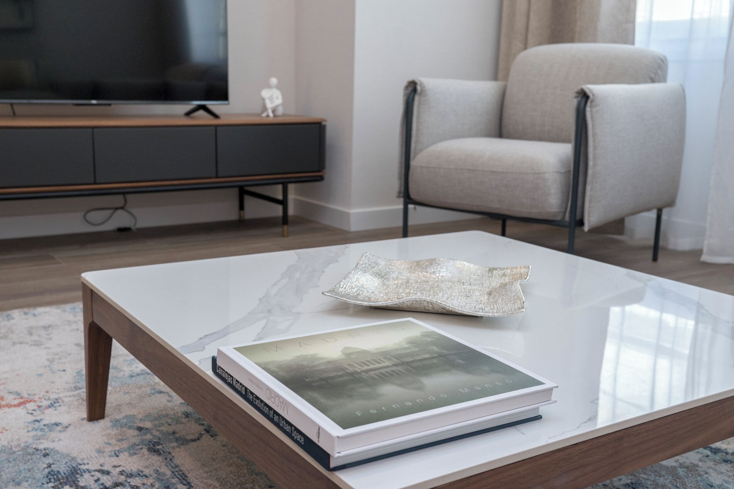 Una sala de estar moderna con una mesa de centro de vidrio, un libro, un televisor, un sofá gris y un mueble de madera.