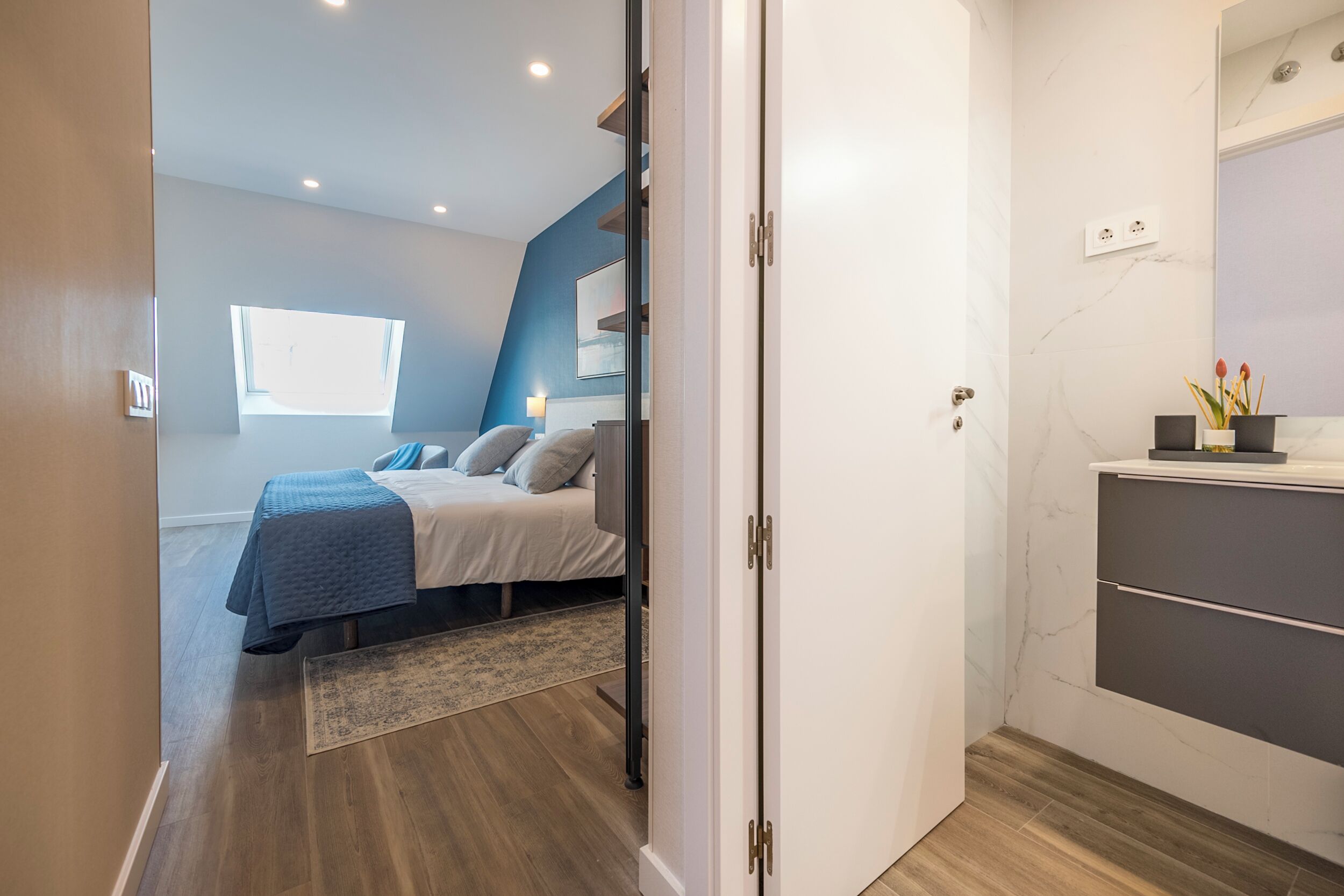 Un dormitorio moderno con cama, mesita de noche, ventana y baño con espejo y lavamanos.