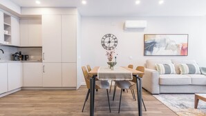 A modern kitchen with white cabinets, a dining table with chairs, a wall clock, and a sofa in a living area. - Apartamentos Dúplex Príncipe de Canalejas (Madrid)