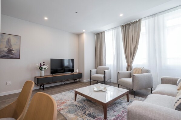 A modern living room with a flat-screen TV, a sofa, armchairs, a coffee table, and a painting on the wall. - Apartamentos Dúplex Príncipe de Canalejas (Madrid)