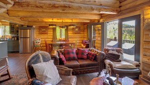 Ein Blockhaushaus-Interieur mit einer Holztreppe, einem Essbereich, einer Couch mit Karo-Kissen und Blick durch große Fenster.