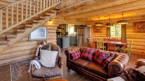 Ein Holzblockhaus-Interieur mit Treppe, einem Wohnzimmer mit Sofa und Sesseln, einem Essbereich und einer Küche.