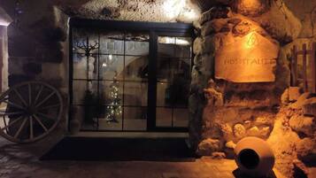 A rustic hotel entrance with a stone wall, a wooden sign reading 'HOTEL ALTO,' and a large wheel.