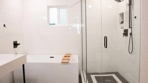 Une salle de bain moderne avec une douche à l’italienne, une baignoire blanche et une petite fenêtre située au-dessus de la baignoire.