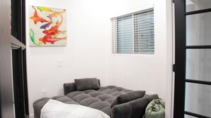 Une pièce avec un canapé gris, un tableau abstrait aux couleurs vives, une fenêtre avec des stores et une porte dotée de panneaux de verre.