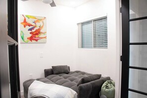 Ein Zimmer mit einem grauen Sofa, einem farbenfrohen abstrakten Gemälde, einem Fenster mit Jalousien und einer Tür mit Glasfeldern.