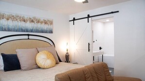 Une chambre à coucher comprenant un lit, une chaise, une table de chevet, une lampe, un tableau et une porte coulissante donnant sur une salle de bain.