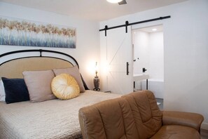 Ein Schlafzimmer mit Bett, Sessel, Nachttisch, Lampe, einem Gemälde und einer Schiebetür zu einem Badezimmer.