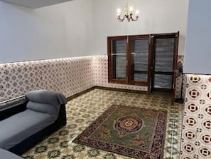 Una habitació amb paper pintat, un sofà, una catifa, una làmpada de cristall i una finestra amb persianes.