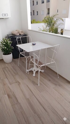 A balcony with a white metal table and chairs, a washing machine, a potted plant, and a view of buildings through a window. - Luxurious Apartment in La Soukra  Comfort Meets Convenience (la soukra)