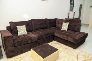 A sectional sofa with patterned cushions, a matching ottoman, and a marble-patterned rug in a room with white walls and a tiled floor. - Cozy 2bed master-ensuite apt in Nairobi near the Airport and SGR train station (Nairobi)